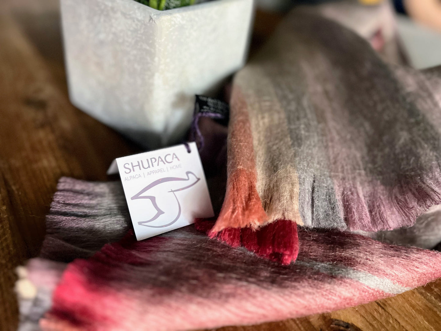A wine and gray Alpaca scarf draped over a wooden surface with a brand tag showing the brand name 'SHUPACA'.