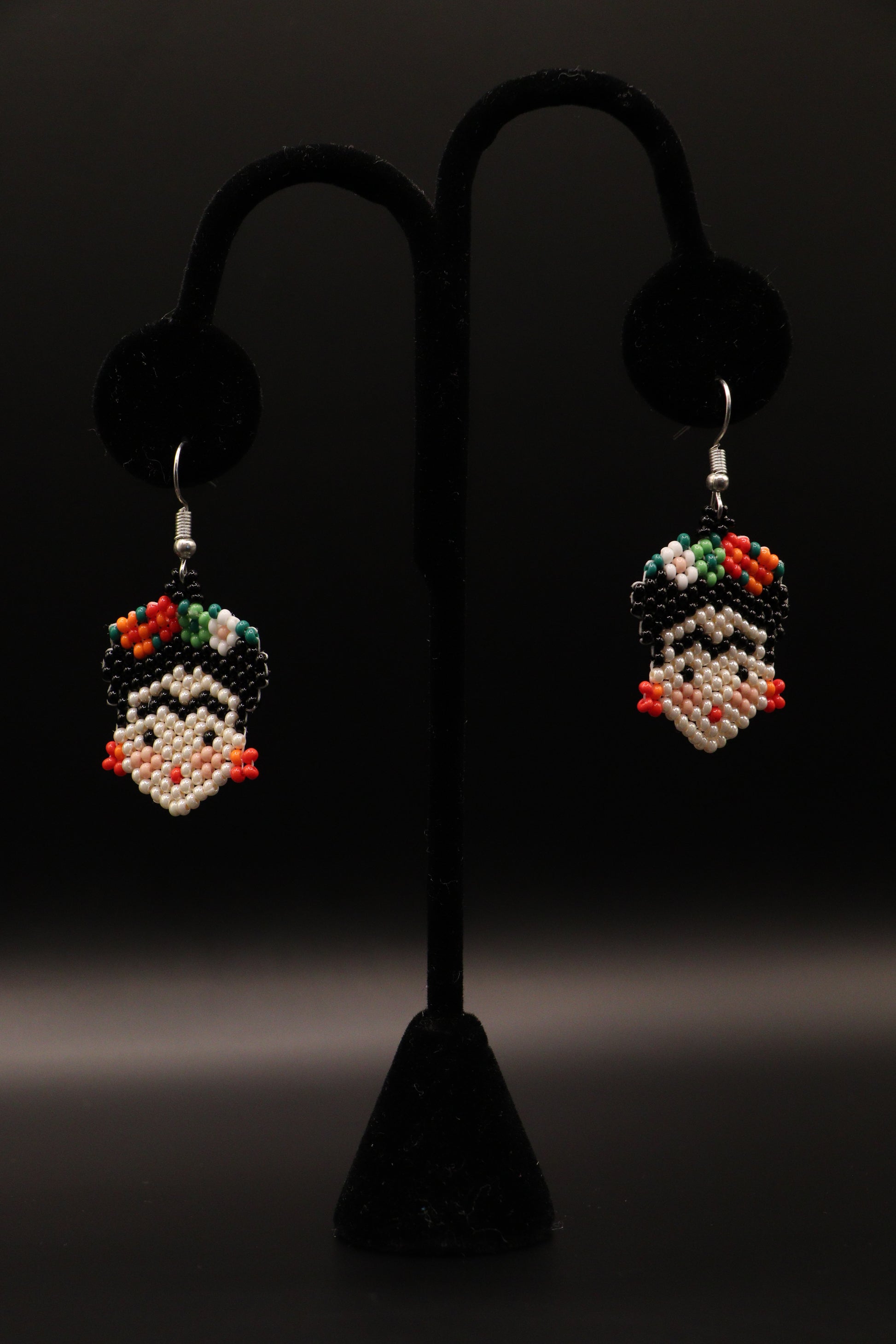 A pair of silver-plated Frida Kahlo inspired earrings featuring intricate seed bead patterns, displayed on a stand against a black background.
