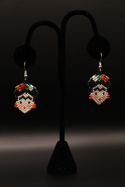 A pair of silver-plated Frida Kahlo inspired earrings featuring intricate seed bead patterns, displayed on a stand against a black background.