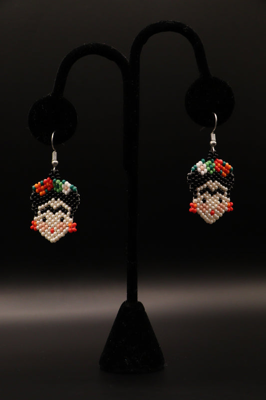 A pair of silver-plated Frida Kahlo inspired earrings featuring intricate seed bead patterns, displayed on a stand against a black background.